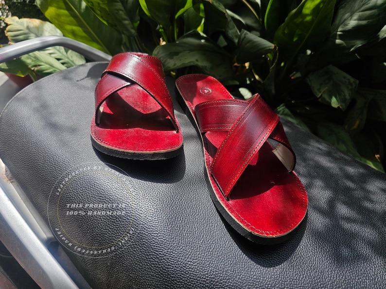 May include: A pair of handmade red leather sandals with a crisscross strap design. The sandals are displayed on a black textured surface, with green foliage in the background. The text "THIS PRODUCT IS 100% HANDMADE" is visible.