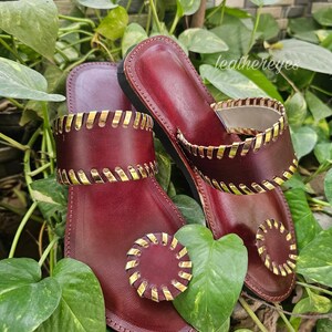 May include: Burgundy leather sandals with gold-coloured accents. The sandals have a thong-style design with a circular embellishment on the toe strap and a decorative band around the ankle. The sandals are surrounded by green foliage.