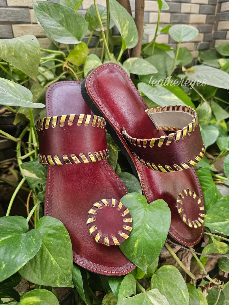 May include: Burgundy leather sandals with gold-coloured accents. The sandals have a thong-style design with a circular embellishment on the toe strap and a decorative band around the ankle. The sandals are surrounded by green foliage.