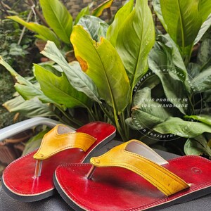Pode incluir: Um par de chinelos de couro feitos &agrave; m&atilde;o. As sand&aacute;lias t&ecirc;m sola vermelha e tira amarela. O texto na imagem diz "THIS PRODUCT IS 100% HANDMADE". O fundo apresenta folhas verdes.