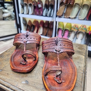 Könnte beinhalten: Ein Paar handgefertigte, braune Ledersandalen mit aufwendigen Ausschnittmustern. Die Sandalen haben eine Zehenschlaufe und einen Knöchelriemen mit dekorativen Nähten und einer strukturierten Oberfläche. Die Sandalen werden auf einer Holzoberfläche präsentiert.