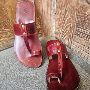 May include: A pair of burgundy leather sandals with a toe loop and a strap across the top, adorned with embossed floral patterns and secured with gold-colored rivets. The sandals are displayed on a textured surface.