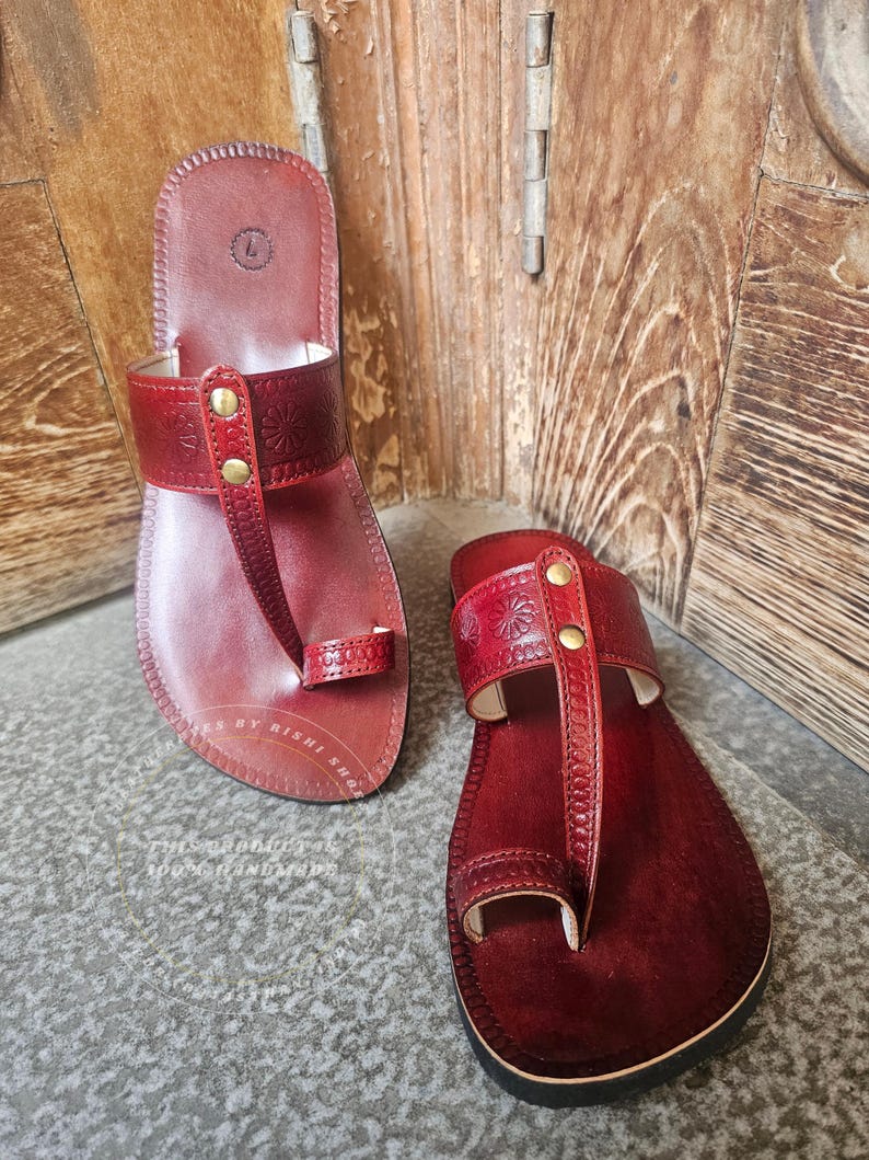 May include: A pair of burgundy leather sandals with a toe loop and a strap across the top, adorned with embossed floral patterns and secured with gold-colored rivets. The sandals are displayed on a textured surface.