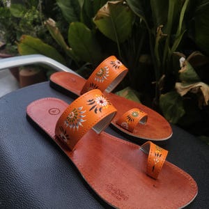 Op de afbeelding: Een paar oranje leren sandalen met bloemmotieven. De sandalen hebben een teengreep en een band over de bovenkant van de voet. De zolen zijn bruin en de sandalen liggen op een zwart oppervlak.