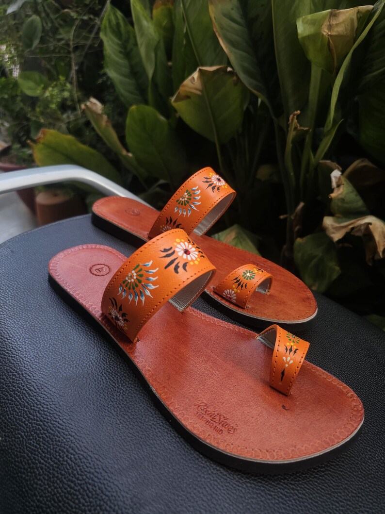 Op de afbeelding: Een paar oranje leren sandalen met bloemmotieven. De sandalen hebben een teengreep en een band over de bovenkant van de voet. De zolen zijn bruin en de sandalen liggen op een zwart oppervlak.