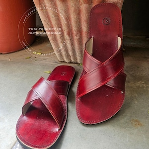 May include: A pair of burgundy leather sandals with a criss-cross strap design. The sandals are handmade and feature a stitched border. The text in the image says "THIS PRODUCT IS 100% HANDMADE".