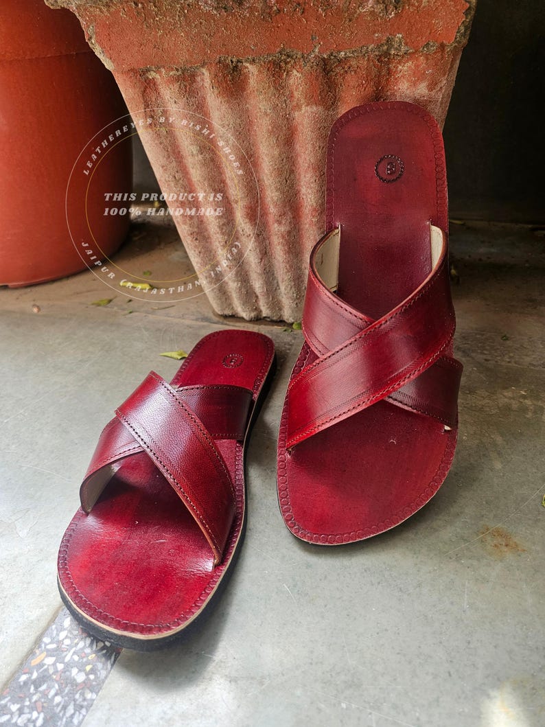 May include: A pair of burgundy leather sandals with a criss-cross strap design. The sandals are handmade and feature a stitched border. The text in the image says "THIS PRODUCT IS 100% HANDMADE".