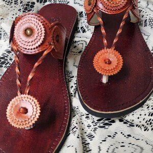 May include: A pair of handcrafted leather sandals. The sandals feature a deep burgundy footbed with a thong strap and decorative floral accents in shades of orange and brown. The sandals are displayed on a white lace surface.