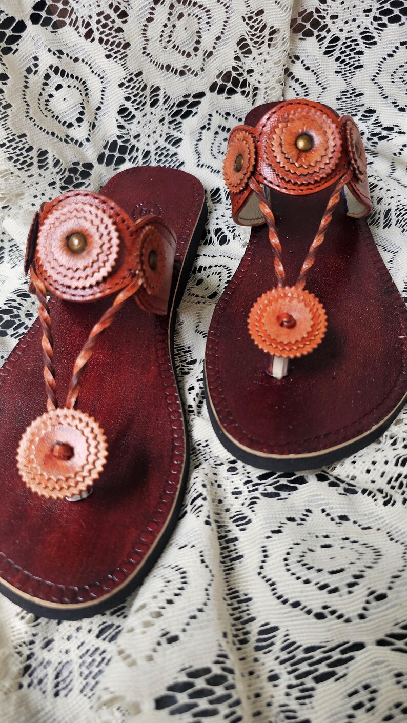 May include: A pair of handcrafted leather sandals. The sandals feature a deep burgundy footbed with a thong strap and decorative floral accents in shades of orange and brown. The sandals are displayed on a white lace surface.