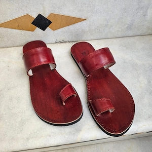 May include: A pair of handcrafted, red leather sandals with a toe loop and a wide strap. The sandals are displayed on a marble surface, showcasing their design and craftsmanship. The sandals are a size 9.