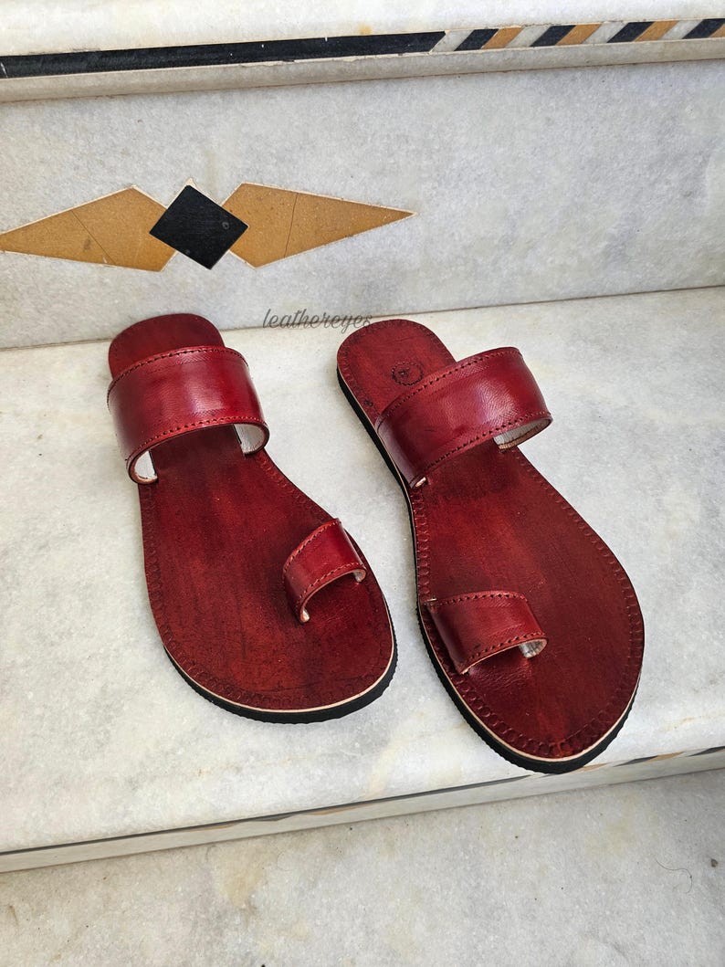 May include: A pair of handcrafted, red leather sandals with a toe loop and a wide strap. The sandals are displayed on a marble surface, showcasing their design and craftsmanship. The sandals are a size 9.