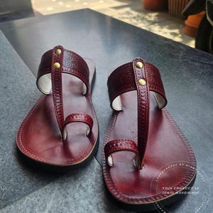 May include: A pair of burgundy leather sandals with a thong-style design. The sandals feature embossed floral patterns and are secured with small, gold-colored rivets. The soles are black, and the sandals appear to be handmade.