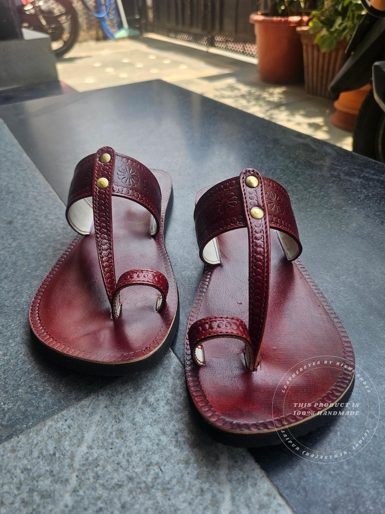 May include: A pair of burgundy leather sandals with a thong-style design. The sandals feature embossed floral patterns and are secured with small, gold-colored rivets. The soles are black, and the sandals appear to be handmade.