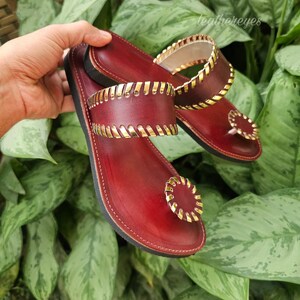 May include: Burgundy leather sandals with gold-coloured accents. The sandals feature a toe ring and straps with a decorative gold trim. The sandals are displayed against a backdrop of green foliage.