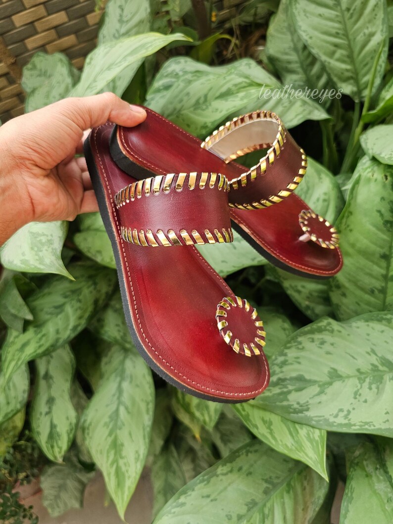 May include: Burgundy leather sandals with gold-coloured accents. The sandals feature a toe ring and straps with a decorative gold trim. The sandals are displayed against a backdrop of green foliage.