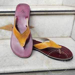 Pode incluir: Um par de chinelos de couro feitos &agrave; m&atilde;o. As palmilhas s&atilde;o de cor bord&ocirc; escuro, com uma tira amarelo-castanho. Os chinelos s&atilde;o tamanho 6. O texto "THERE EYES BY RISHI SHOES" e "THIS PRODUCT IS 100% HANDMADE" s&atilde;o vis&iacute;veis.