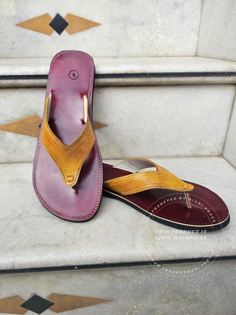 Pode incluir: Um par de chinelos de couro feitos &agrave; m&atilde;o. As palmilhas s&atilde;o de cor bord&ocirc; escuro, com uma tira amarelo-castanho. Os chinelos s&atilde;o tamanho 6. O texto "THERE EYES BY RISHI SHOES" e "THIS PRODUCT IS 100% HANDMADE" s&atilde;o vis&iacute;veis.