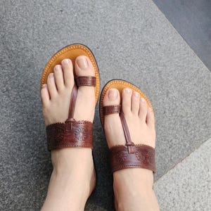 May include: A pair of brown leather sandals featuring a toe-post design and a wide horizontal strap across the midfoot. The strap is decorated with an embossed floral pattern and scalloped edges. The footbed is tan with a stitched border.