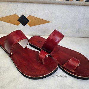 May include: A pair of burgundy leather sandals with a toe loop and a strap across the top. The sandals have a dark sole and a white interior. The sandals are displayed on a marble surface. The text "leathereyes" is visible.