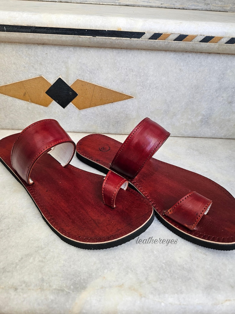 May include: A pair of burgundy leather sandals with a toe loop and a strap across the top. The sandals have a dark sole and a white interior. The sandals are displayed on a marble surface. The text "leathereyes" is visible.