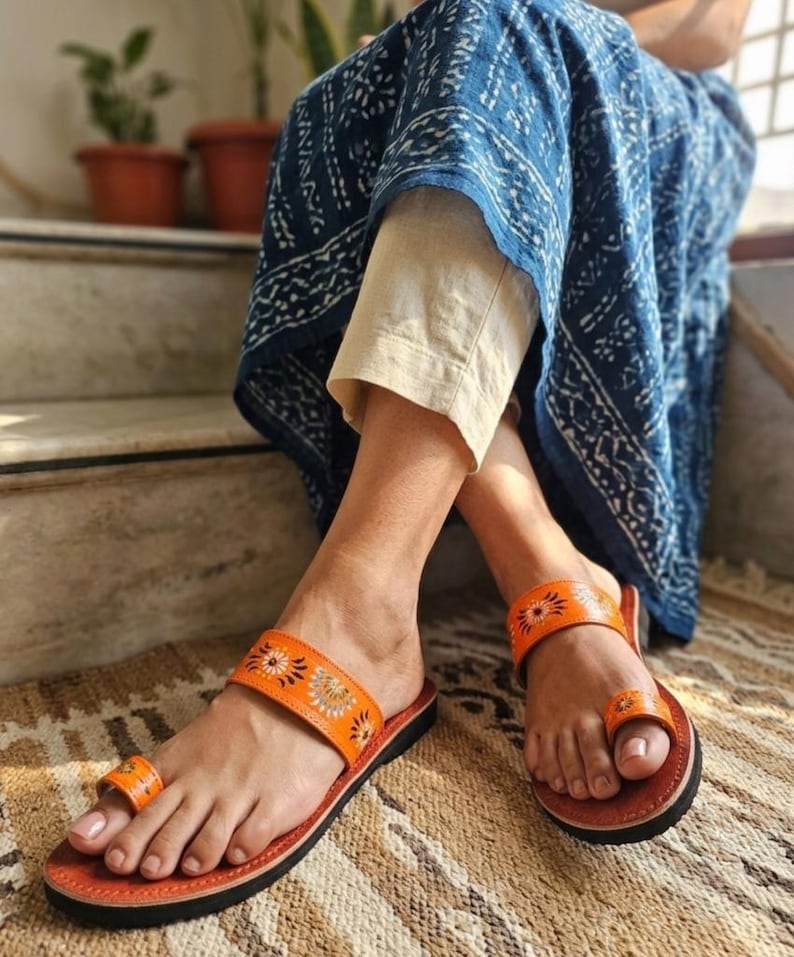 Op de afbeelding: Oranje leren sandalen met bloemenborduurwerk. De sandalen hebben een teengreep en een band over de bovenkant van de voet. De zolen zijn zwart en het voetbed is bruin. De sandalen liggen op een patroonkleed.