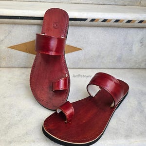 May include: A pair of red leather sandals with a toe loop and a wide strap across the top of the foot. The sandals have a black sole and are displayed on a marble surface. The sandals are handmade and have a classic design.