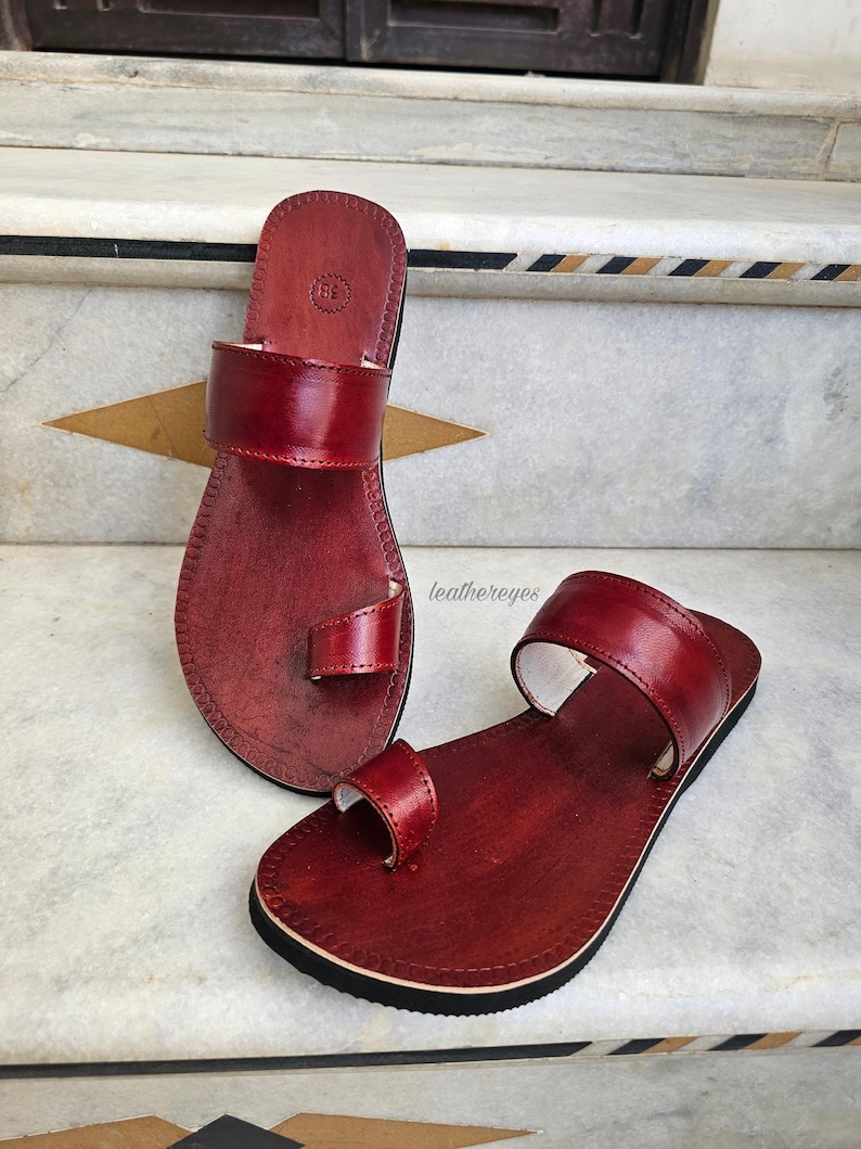May include: A pair of red leather sandals with a toe loop and a wide strap across the top of the foot. The sandals have a black sole and are displayed on a marble surface. The sandals are handmade and have a classic design.