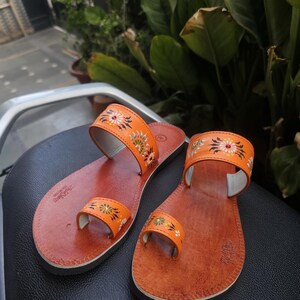 Op de afbeelding: Een paar oranje leren sandalen met bloemmotieven. De sandalen hebben een teengreep en een band over de bovenkant van de voet, beide versierd met kleurrijke bloempatronen. De zolen zijn een donkerdere bruintint.