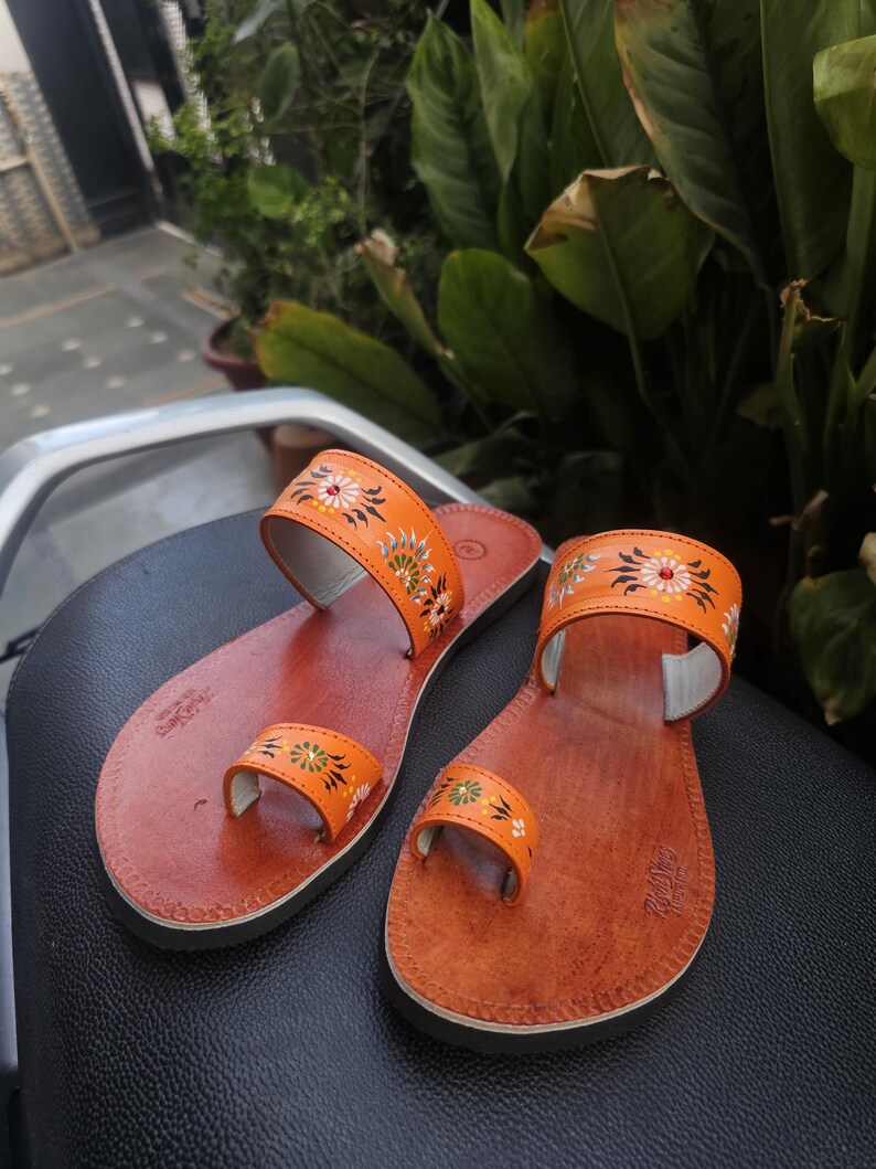 Op de afbeelding: Een paar oranje leren sandalen met bloemmotieven. De sandalen hebben een teengreep en een band over de bovenkant van de voet, beide versierd met kleurrijke bloempatronen. De zolen zijn een donkerdere bruintint.