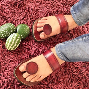 May include: A pair of burgundy leather sandals with a toe loop and ankle strap. The sandals are worn on a burgundy shag rug. Decorative green painted eggs with white patterns are in the background.