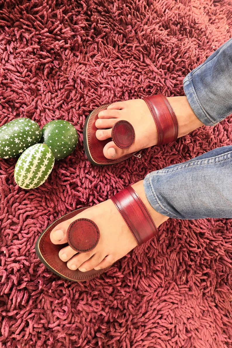 May include: A pair of burgundy leather sandals with a toe loop and ankle strap. The sandals are worn on a burgundy shag rug. Decorative green painted eggs with white patterns are in the background.