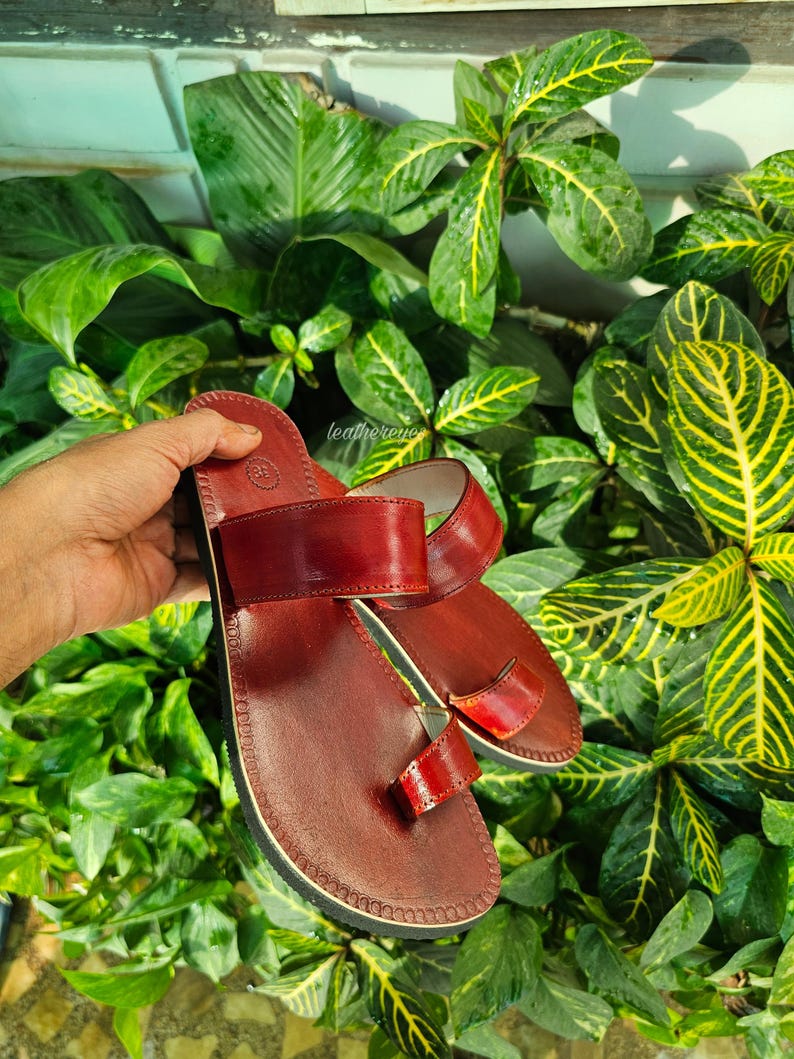 May include: A pair of burgundy leather sandals with a toe loop and two straps. The sandals are set against a backdrop of lush green foliage. The brand name "leathereye" is visible on the side.