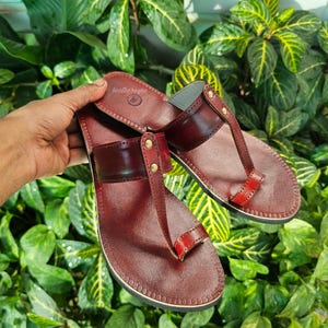 May include: Burgundy leather sandals with a toe loop and a wide strap across the top. The sandals have a stitched sole and are adorned with small gold-colored rivets. The brand name "leathereges" is visible on the top of the sandal.