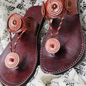 May include: A pair of handcrafted leather sandals in a rich burgundy hue. The sandals feature decorative, circular accents in a lighter shade of brown, and a braided thong strap. The sandals are displayed on a white lace surface.
