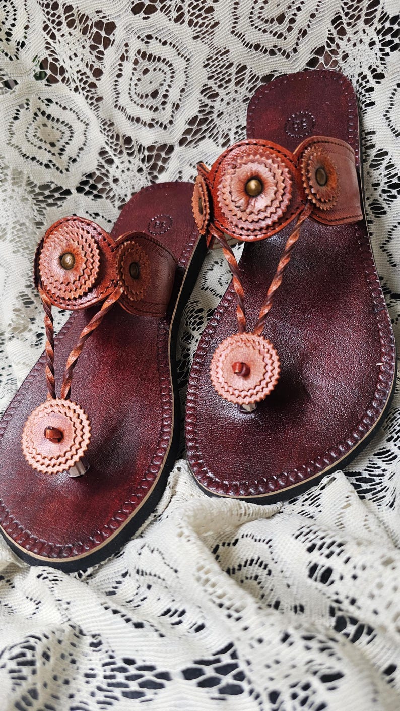 May include: A pair of handcrafted leather sandals in a rich burgundy hue. The sandals feature decorative, circular accents in a lighter shade of brown, and a braided thong strap. The sandals are displayed on a white lace surface.