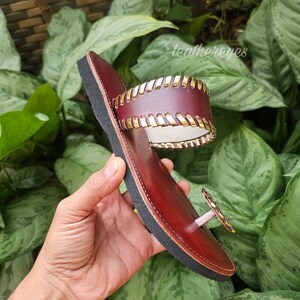 May include: Burgundy leather sandal with a black sole and gold-coloured detailing. The sandal has a toe post and a wide strap with gold stitching. The brand name "leathereyes" is visible in the background.