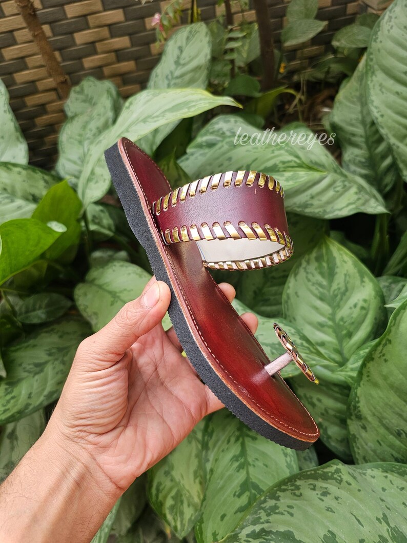 May include: Burgundy leather sandal with a black sole and gold-coloured detailing. The sandal has a toe post and a wide strap with gold stitching. The brand name "leathereyes" is visible in the background.