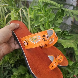 Op de afbeelding: Oranje leren sandalen met geschilderde bloemmotieven. De sandalen hebben een teensluiting en een band over de voet, versierd met kleurrijke bloemdesigns. De sandalen staan tegen een achtergrond van groen gebladerte.