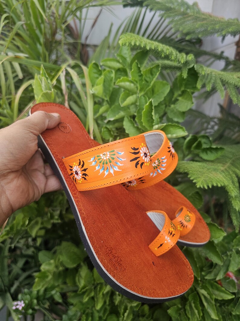 Op de afbeelding: Oranje leren sandalen met geschilderde bloemmotieven. De sandalen hebben een teensluiting en een band over de voet, versierd met kleurrijke bloemdesigns. De sandalen staan tegen een achtergrond van groen gebladerte.