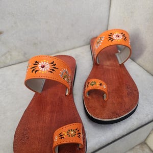 Op de afbeelding: Een paar oranje leren sandalen met bloemenborduurwerk. De sandalen hebben een teengreep en een band over de bovenkant van de voet. De zolen zijn bruin met een zwarte rand. De sandalen zijn handgemaakt.