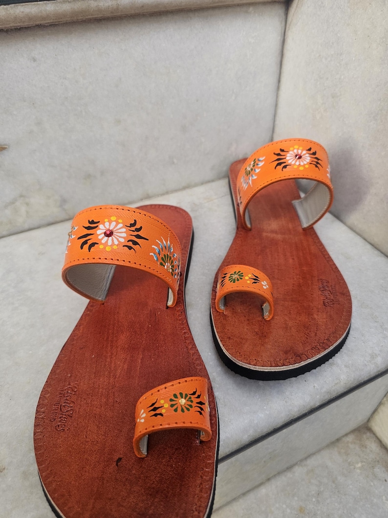 Op de afbeelding: Een paar oranje leren sandalen met bloemenborduurwerk. De sandalen hebben een teengreep en een band over de bovenkant van de voet. De zolen zijn bruin met een zwarte rand. De sandalen zijn handgemaakt.