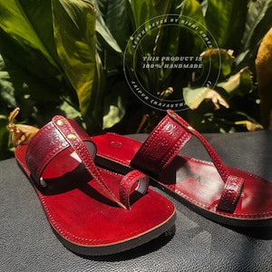 May include: A pair of handmade, red leather sandals with floral embossed detailing. The sandals feature a toe loop and a strap across the top of the foot, secured with small gold-colored studs. The text "THIS PRODUCT IS 100% HANDMADE" is visible.