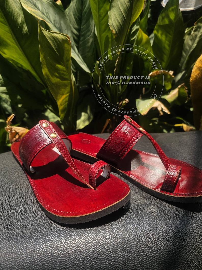 May include: A pair of handmade, red leather sandals with floral embossed detailing. The sandals feature a toe loop and a strap across the top of the foot, secured with small gold-colored studs. The text "THIS PRODUCT IS 100% HANDMADE" is visible.