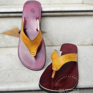 Pode incluir: Um par de sand&aacute;lias de couro feitas &agrave; m&atilde;o. As sand&aacute;lias t&ecirc;m uma base bord&ocirc; escura com uma tira amarela. A sola &eacute; preta. As sand&aacute;lias s&atilde;o tamanho 6. O texto "LEATHEREYES BY RISHI SHOES" e "THIS PRODUCT IS 100% HANDMADE" s&atilde;o vis&iacute;veis.