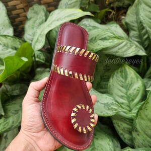 May include: Burgundy leather sandal with gold-coloured accents. The sandal features a toe loop and a strap across the top of the foot, both adorned with gold detailing. The text "leathereyes" is visible on the side.