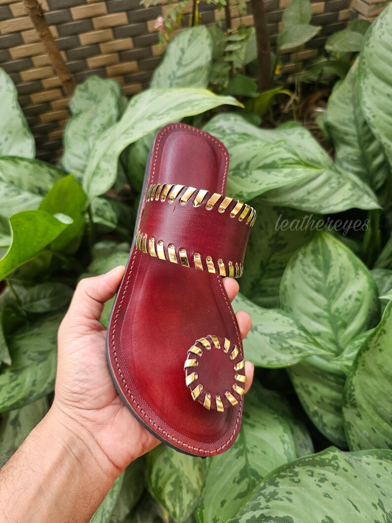 May include: Burgundy leather sandal with gold-coloured accents. The sandal features a toe loop and a strap across the top of the foot, both adorned with gold detailing. The text "leathereyes" is visible on the side.