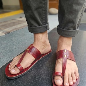 May include: Red leather sandals with a toe loop and a strap across the top of the foot. The sandals have a decorative floral pattern and are secured with small gold-colored rivets. The soles are dark brown.