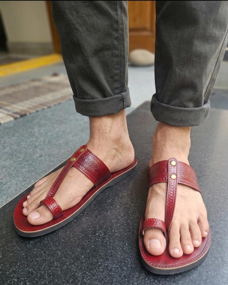 May include: Red leather sandals with a toe loop and a strap across the top of the foot. The sandals have a decorative floral pattern and are secured with small gold-colored rivets. The soles are dark brown.