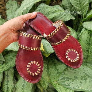 May include: Burgundy leather sandals with gold-coloured accents. The sandals feature a decorative gold trim around the edges and straps. The design includes a circular gold embellishment on the toe area. The sandals are displayed against a backdrop of green foliage.