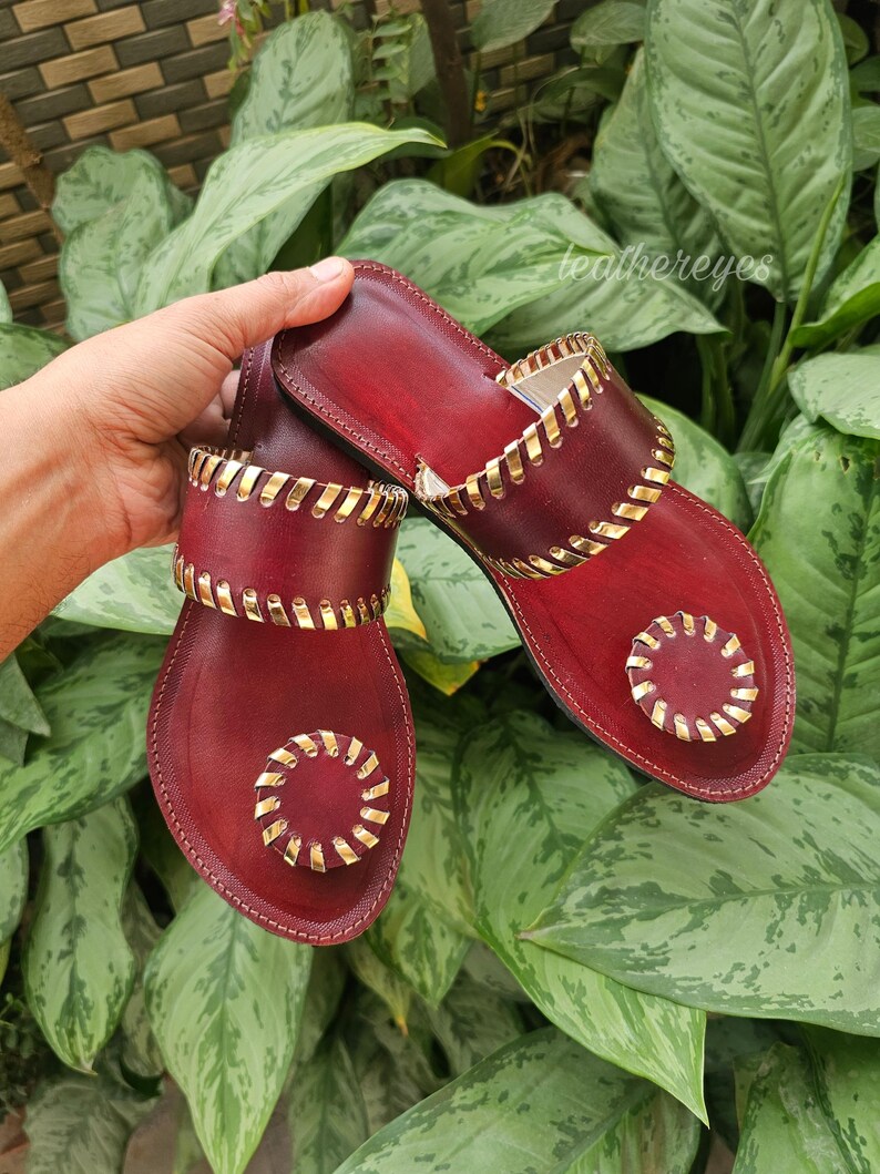 May include: Burgundy leather sandals with gold-coloured accents. The sandals feature a decorative gold trim around the edges and straps. The design includes a circular gold embellishment on the toe area. The sandals are displayed against a backdrop of green foliage.